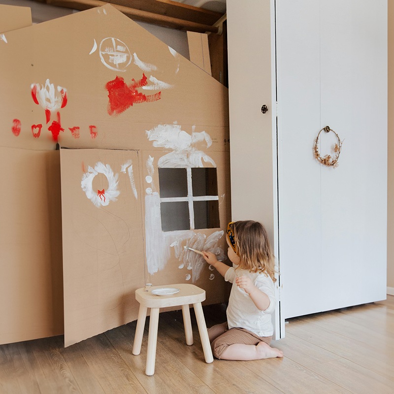 Kind mit Hocker malt auf einem Kartonhaus